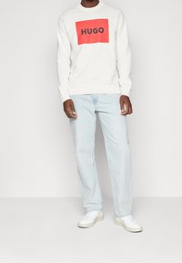 Felpa bianca con una grafica rettangolare rossa che presenta "HUGO" in nero. Abbinata a jeans in denim azzurro chiaro e sneakers bianche.