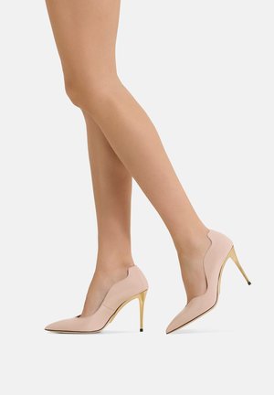 Gambe con décolleté rosa chiaro a punta con tacchi a spillo dorati, un piede leggermente sollevato, su uno sfondo chiaro uniforme.