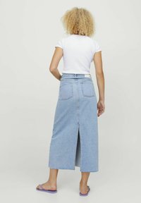 Denim maxi skirt in light blue, featuring a center slit at the back, two rear pockets, and a raw hem. Paired with a white fitted top.
