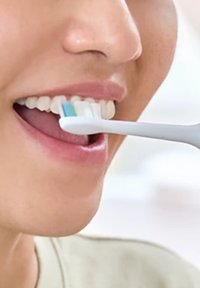 Spazzolino da denti bianco con setole blu posizionato vicino ai denti, focalizzandosi sul movimento di spazzolamento; pelle liscia e gengive visibili in primo piano.