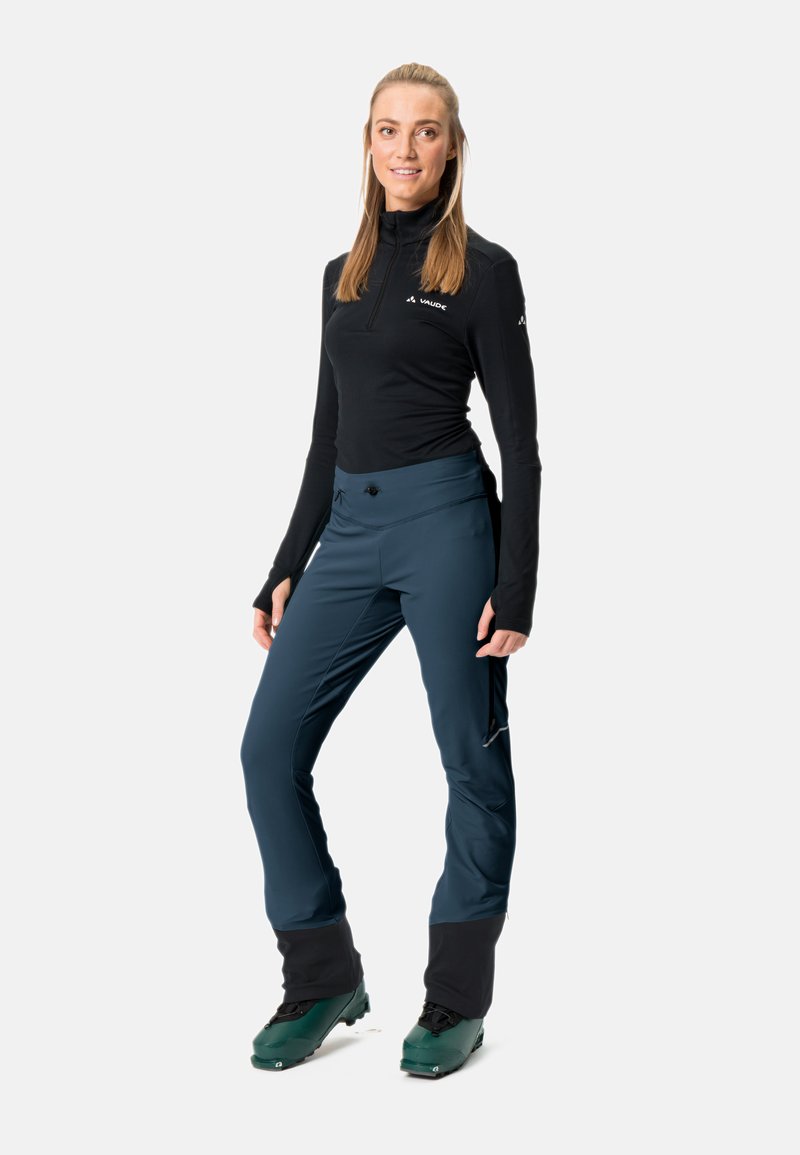 Schwarzes Langarmoberteil und marineblaue Skihosen mit Seitentaschen, kombiniert mit grünen Skischuhen. Die Hose hat schmal zulaufende Beine und verstellbare Bündchen.