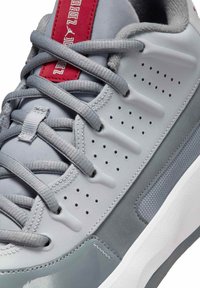 Zbliżenie na szary tenisówkę z perforowanymi bocznymi panelami, szarymi sznurowadłami, czerwonym językiem i teksturowaną białą i szarą podeszwą.