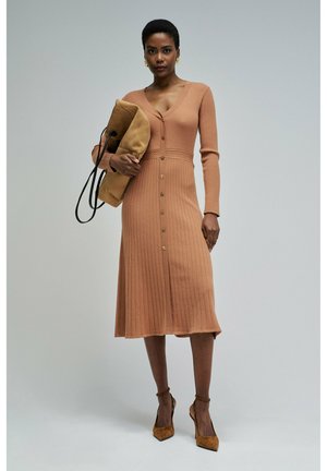 Mujer con un vestido midi de manga larga, abotonado y acanalado en color camel, sosteniendo un bolso grande color marrón claro, con zapatos de tacón altos marrones a juego y pendientes dorados.