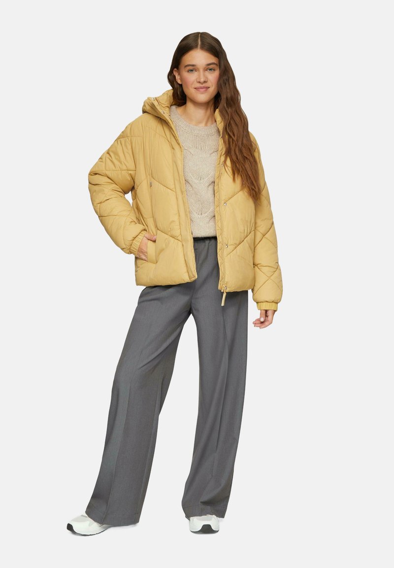 Geel gewatteerde pufferjack met capuchon, beige gebreide trui en grijze wijde pantalon. Model draagt witte sneakers.