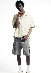 Veste beige oversize zippée à manches courtes, associée à un short cargo gris avec poches latérales et détail en chaîne, portée avec des chaussettes blanches et des baskets.