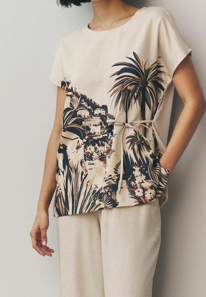 Haut beige à manches courtes avec imprimé paysage tropical et ceinture à la taille, associé à un pantalon beige ample, porté par une personne aux cheveux foncés.
