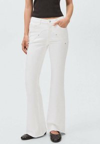 Jeans acampanados blancos de mezclilla con bolsillos frontales; presenta un detalle de costura curva y acentos de herrajes en tono plateado; el dobladillo está deshilachado.
