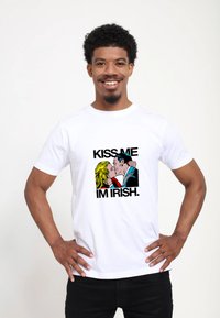 Bílá tričko s grafickým designem páru, který se líbá, a nápisem "KISS ME I'M IRISH" tučným černým písmem v barevném karikaturním stylu.