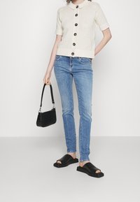 Stickad kortärmad cardigan i krämfärg med stora knappar, tillsammans med ljusblå jeans och svarta slidesandaler. Bär en liten svart väska.