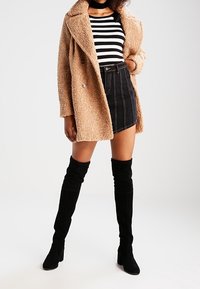 Manteau beige duveteux à double boutonnage porté sur un haut rayé noir et blanc, assorti à une jupe en jean noire et des bottes noires montantes.