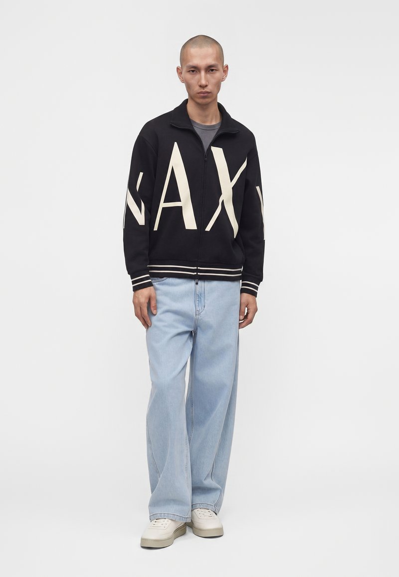 Homme portant une veste noire à fermeture éclair avec de grandes lettres blanches, un jean large bleu clair et des baskets blanches, debout devant un fond uni.