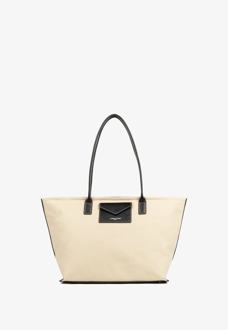 Beige canvas boodschappentas met zwarte leren accenten, vlakke bodem en lange hengsels. Heeft een zwarte pocket met stikdetails aan de voorkant.
