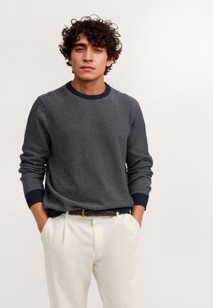 Młody mężczyzna z kręconymi włosami, ubrany w ciemny wzorzysty sweter i białe spodnie, stoi z rękami w kieszeniach na jednolitym tle.