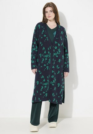 Un long cardigan navy avec un motif floral vert, à front ouvert et avec des fentes latérales, associé à un haut vert et un pantalon sombre. Texture en tricot douce.