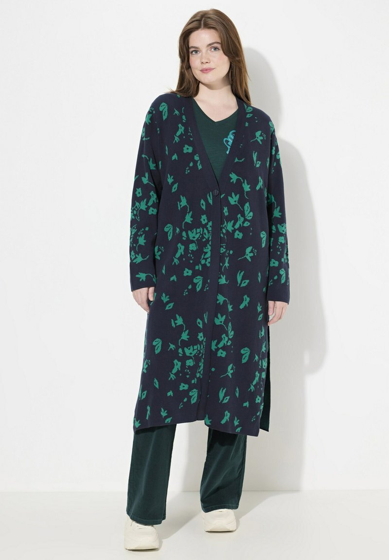 Lång marinblå kofta med grönt blommönster, öppen framtill och sidsprund, kombinerad med en grön topp och mörka byxor. Mjuk stickad textur.
