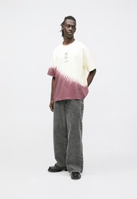 Lühikeste varrukatega t-särk kreemikas toonis, mis on maroonse dip-dye efektiga ja millel on rinnal graafiline trükk. Sobitatud lahtiste mustade teksadega.