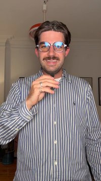 Blå og hvitstripete skjorte med knapper, runde blå briller med reflekterende glass, holder en mikrofon, smilende, innendørs miljø.