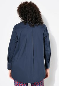 Navy-blaues Hemd mit langen Ärmeln, geknöpften Manschetten und einem Rückenfalten-Detail. Getragen mit gemusterten roten Hosen mit blauen Akzenten.