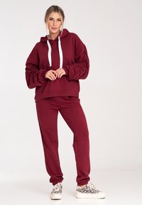 Burgundowa bluza z kapturem z białymi sznurkami, ściągaczami na rękawach i kieszenią; w zestawie z pasującymi spodniami dresowymi i wzorzystymi trampkami.