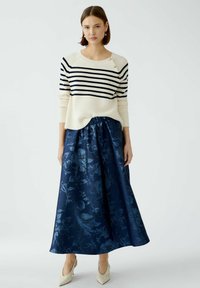 Gestreifter cremefarbener und marineblauer Pullover mit Knopfdetail, kombiniert mit einem marineblau gemusterten Maxirock, ergänzt durch beige High Heels.