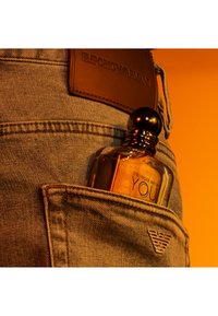 Klart glass parfymeflaske med svart lokk, merket "Stronger With You", delvis i en denimlomme, mot en oransje bakgrunn.