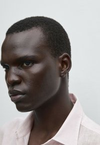 Side profile of a person with dark skin, short, textured hair, wearing a soft pink collared shirt against a plain light background.