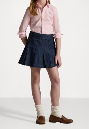 Dítě má na sobě růžovou košili na knoflíky zastrčenou do tmavé džínové plisované sukně, bílé ponožky a hnědé mokasíny, stojí se založenýma rukama za zády.