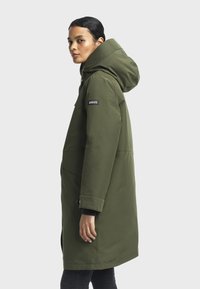 Parka à capuche vert olive avec fermeture avant zippée et à boutons-pression, comportant un logo sur la manche et une coupe droite.
