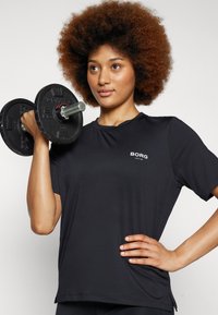Svart atletisk t-skjorte med korte ermer, med en liten hvit logo på brystet og en glatt, lett stofftekstur.