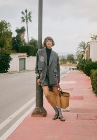 Γυναίκα με γκρι σακάκι με ρίγες και σορτς στηρίζεται σε δρόμο με φανάρι, κρατώντας πλεκτό καλάθι, στέκεται σε ροζ πεζοδρόμιο με φοινικόδεντρα στο φόντο.