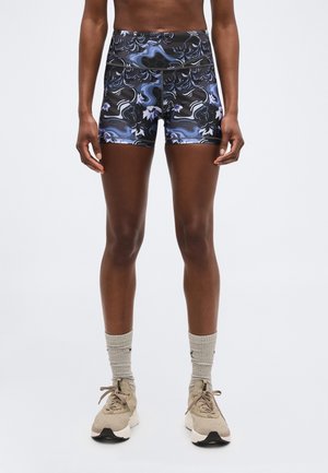 Αθλητικό άτομο που φοράει ποδηλατικά σορτς υψηλής μέσης με μοτίβο μωβ και μαύρο, μπεζ κάλτσες και μπεζ αθλητικά παπούτσια, στέκεται μπροστά από μια απλή φωτεινή επιφάνεια.