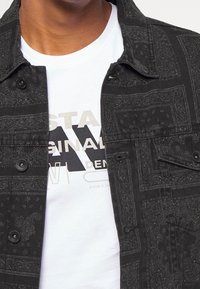 Veste en denim noir avec texture à motifs, fermeture à boutons sur le devant et accents détaillés. T-shirt blanc avec imprimé en dessous.