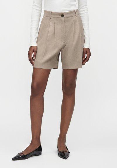 Femme portant un short beige taille haute sur mesure avec des plis, un haut blanc à manches longues, et des ballerines noires pointues, debout devant un fond blanc.