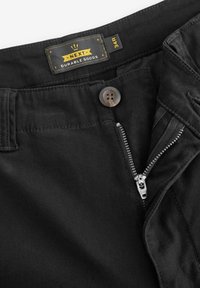 Schwarze Baumwollhosen mit glatter Textur, ausgestattet mit einem Metallreißverschluss, Knopfverschluss und einem Etikett mit der Aufschrift "NEXT DURABLE GOODS". Größe 34R.