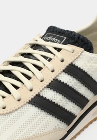 Κοντινή λήψη ενός μπεζ και μαύρου αθλητικού παπουτσιού Adidas με κρεμ κορδόνια, που δείχνει υφή υφάσματος, ραφές σε ζιγκ-ζαγκ και το λογότυπο της Adidas στη γλώσσα.