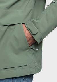 Giacca impermeabile verde con tasca frontale, orlo e polsini regolabili, texture liscia e cordino nascosto all'orlo.