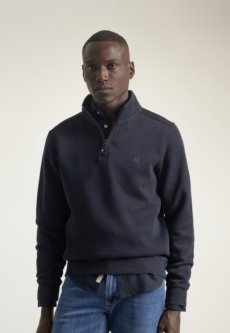 Pulóver de media cremallera azul marino, hecho de un tejido texturizado con puños y dobladillo acanalados. Detalle del logo notable en el pecho, superpuesto a una camisa con cuello.