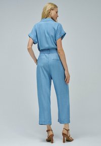 Mono de chambray azul claro con mangas cortas enrolladas, cintura ceñida con cinturón y diseño de pierna ancha. Presenta costuras sutiles y bolsillos traseros.