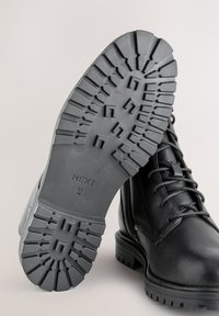 Zwarte leren laars met een dikke rubberen zool voorzien van een robuust profiel. Zij- en voorkant tonen vetersluiting en stikdetails.