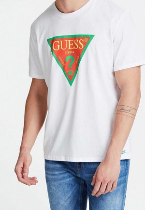 Homme portant un t-shirt blanc à manches courtes avec le logo rouge et vert Guess Lisboa et un jean bleu, montrant un tatouage sur l'avant-bras gauche.