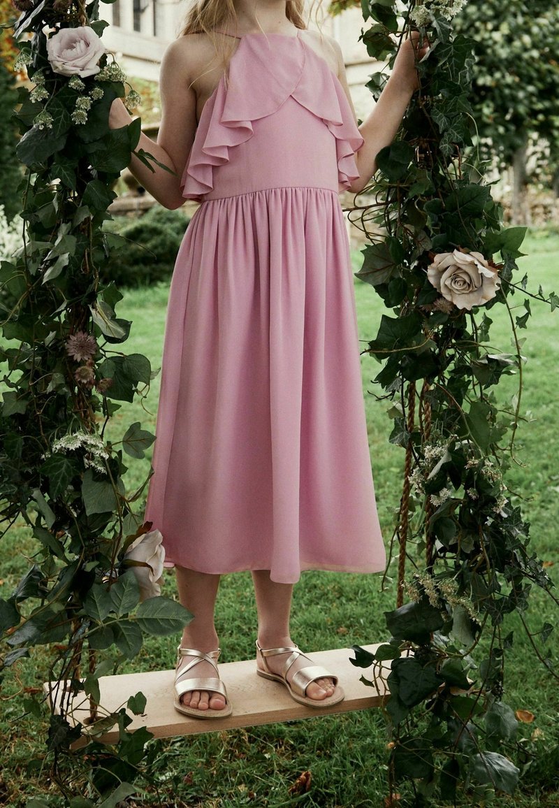 Enfant en robe rose à volants et sandales dorées debout sur une balançoire en bois décorée de feuilles vertes et de roses claires dans le jardin.
