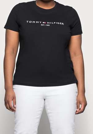 Person trägt ein schwarzes Tommy Hilfiger T-Shirt mit Logo und dem Schriftzug "EST. 1985", kombiniert mit weißen Hosen vor einem schlichten Hintergrund.
