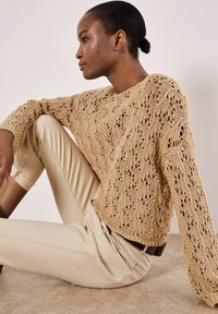 Beige gestrickter Pullover mit offenem Spitzenmuster und langen Ärmeln, kombiniert mit cremefarbenen, figurbetonten Hosen. Getragen mit einem braunen Gürtel.