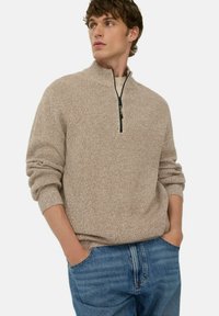 Beige gebreide trui met een hoge kraag en een halve rits. Geribde manchetten en zoom, met een textuurpatroon. Gecombineerd met blauwe jeans.