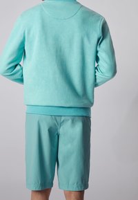 Persona che indossa una felpa turchese e pantaloni corti abbinati fino al ginocchio, vista da dietro su uno sfondo grigio chiaro uniforme.