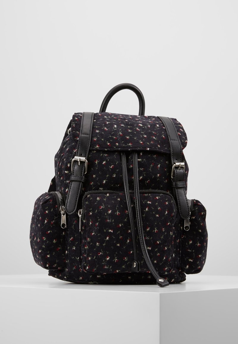 Sac à dos en tissu noir avec petit motif floral rouge et blanc, poches zippées avant et latérales, bretelles en cuir et poignée supérieure sur surface blanche.