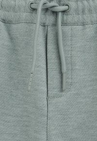 Nahaufnahme eines hellgrauen Stoffbunds mit einer durch Metallösen gefädelten Kordel.