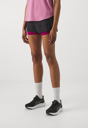 Schwarze Sportshorts mit pinken Akzenten, kombiniert mit einem hellpinkfarbenen Shirt und schwarzen Laufschuhen, die von weißen Knöchelsocken begleitet werden.
