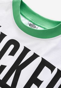 Bijela majica s podebljanim crnim natpisom, okruglim zelenim rebrastim ovratnikom i oznakom s "wicked" logom na unutarnjoj strani vrata.