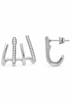 Paire de boucles d’oreilles créoles polies, de ton argenté, aux courbes ouvertes et sculpturales, ornées de rangées de pierres transparentes sur un côté. Une boucle est présentée de face et l’autre de profil.